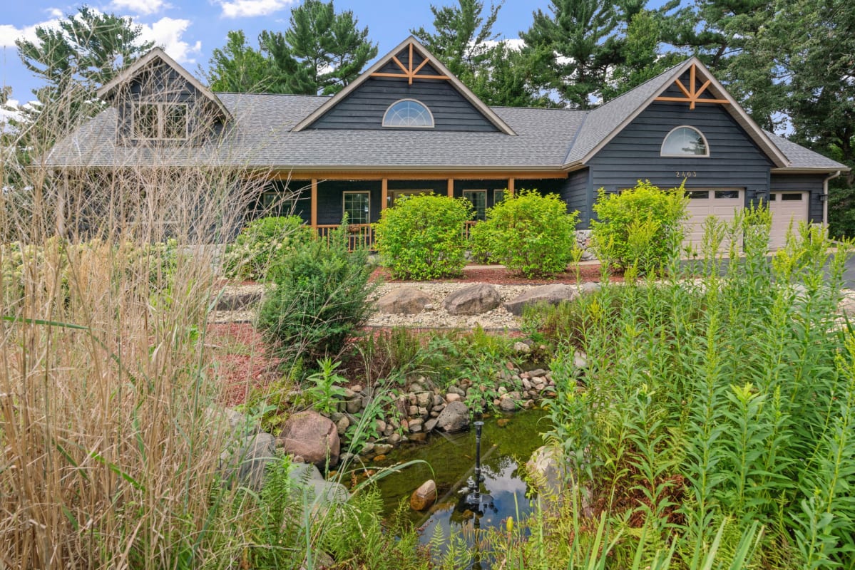 Charming lakeside retreat nestled among towering pines with beautifully landscaped gardens and tranquil water feature at the entrance.