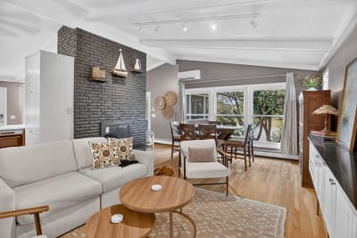 A cozy open-concept living and dining space featuring a black brick fireplace, white ceiling beams, and large windows showcasing greenery outside.