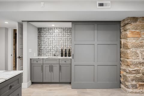 A modern private bar area featuring gray cabinetry, marble counters, and a textured silver tile backsplash.