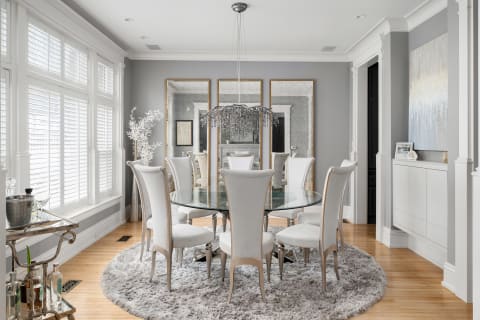 A sophisticated dining room featuring a glass table, plush chairs, and a crystal chandelier under natural light from large windows.