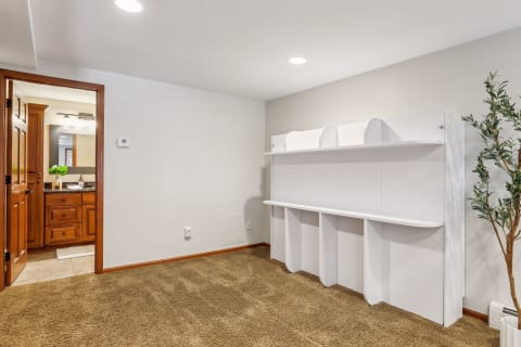 A cozy room with white storage shelving, beige carpet, and a small plant, leading to a wooden bathroom door.