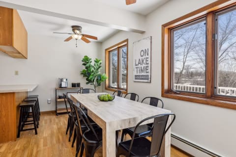A cozy dining room with a handcrafted wood table, metal chairs, large windows, and a sign that reads 'ON LAKE TIME'.