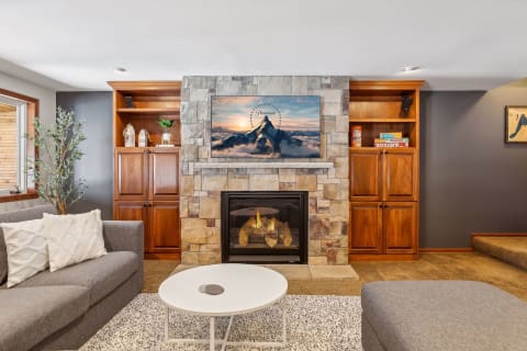 A cozy living room with a stone fireplace, plush gray sofas, and a large mountain scene displayed on the TV.