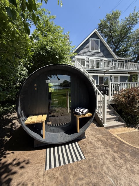 A barrel-shaped sauna with glass doors in a garden, featuring wooden benches and a striped rug, with a house in the background.