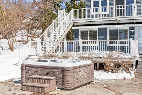 A hot tub on a snowy patio near a house with stair access and outdoor deck space.