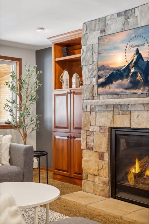 Cozy living room featuring a stone fireplace, wooden cabinetry, plush seating, and a large TV displaying mountain scenery.