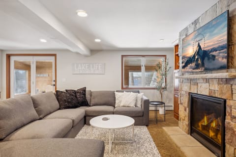 A cozy living room with a gray sectional sofa, natural stone fireplace, and a mountain scene on the TV.
