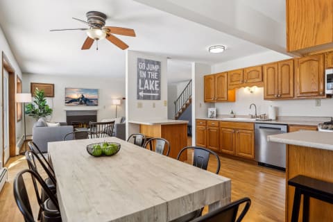 A bright dining area with a wooden table, black chairs, and a cozy living room, showcasing a kitchen and decorative wall art.