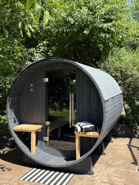 Barrel-style sauna set in a green landscape with wooden shelves and a striped mat at the entrance.