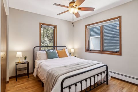 A cozy bedroom featuring a metal bed with white linens and colorful pillows, soft lighting from two bedside lamps, and windows providing natural light.