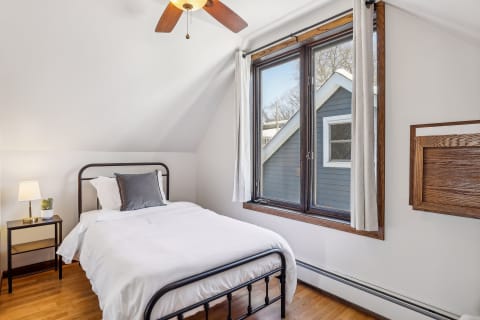 A cozy bedroom featuring a single bed, large window, and minimalist decor with wooden accents.