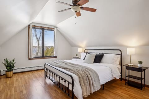 A cozy upstairs bedroom with vaulted ceilings, a large window with a lake view, and stylish decor including a plush bed and green plant.