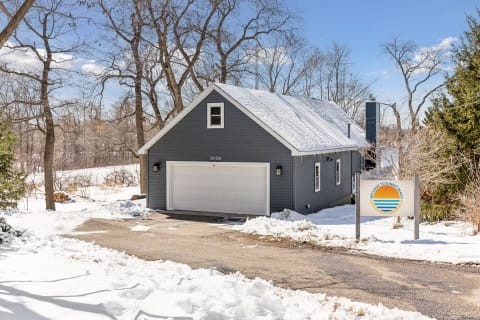 A gray vacation home in winter, surrounded by snow and trees, featuring a welcoming sign.