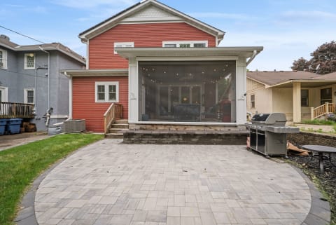 Spacious paver patio with a red house in the background, featuring string lights and an outdoor grill.