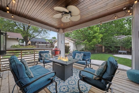 A covered patio featuring comfortable blue seating, a central fire table, string lights, and a glimpse of a verdant backyard.