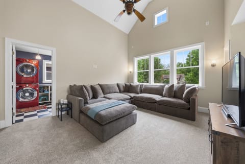 A spacious living room featuring a plush sectional sofa, large windows, and a laundry area with red appliances visible in the background.