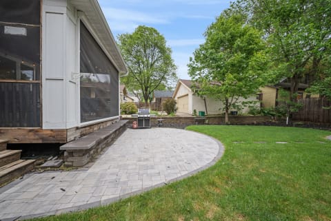A spacious paved patio with a grill, surrounded by a green lawn and trees in a private backyard.