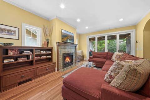A warm living room with a rust-red sectional sofa, a fireplace, bookshelves, and garden views through glass doors.