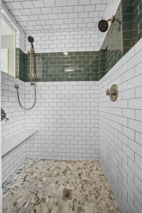 A luxurious shower featuring white subway tiles, a green accent band, dual showerheads, and a built-in bench, complete with a unique pebble floor.