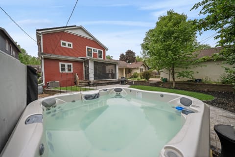 A tranquil backyard with a hot tub, red house, and mature trees under a clear sky.
