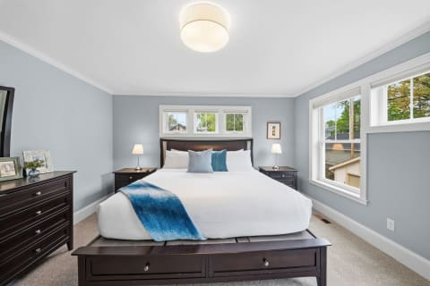 A tranquil bedroom featuring blue-gray walls, a king-size bed with white linens, and soft natural lighting from large windows.
