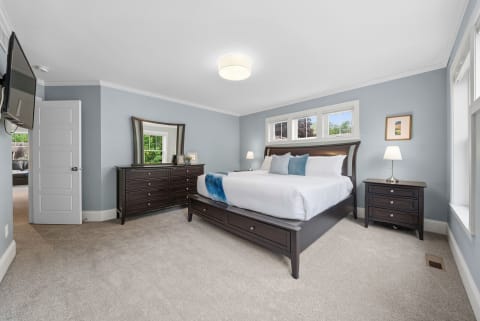 A cozy bedroom featuring soft blue walls, a comfortable bed with crisp linens, natural light, and elegant wooden furniture.