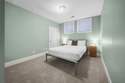 A brightly lit bedroom featuring sage green walls, a neatly made bed, nightstands, and soft carpeting.
