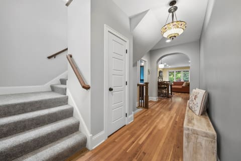 A warm hallway with hardwood floors leading to a dining space, featuring a chandelier and carpeted stairs.