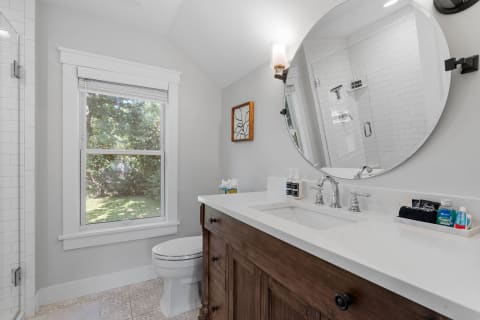 A bright modern bathroom featuring a circular mirror, double sink vanity, and a view of greenery through the window.