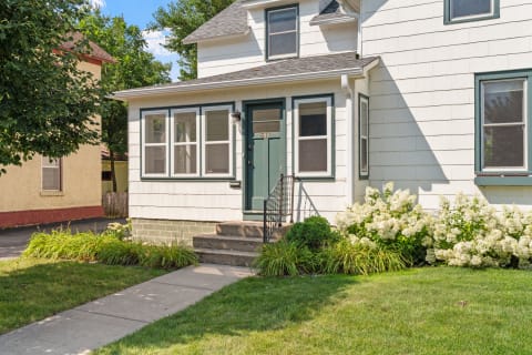 Front porch of a two-story house with white siding, green trim, and blooming flowers.