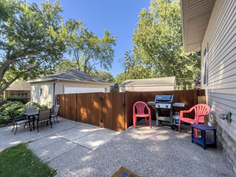 Outdoor space with a wooden fence, pink chairs, a grill, and a dining table in a green setting.