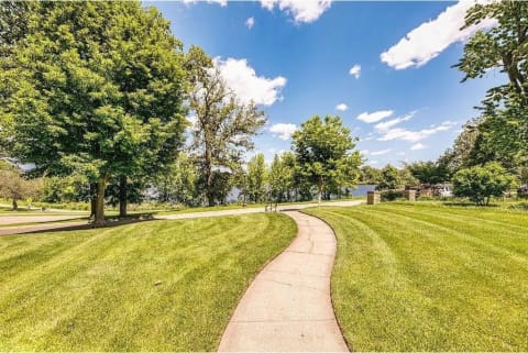 A curved concrete path through well-kept grass and trees, leading to a distant lake under a blue sky with clouds.