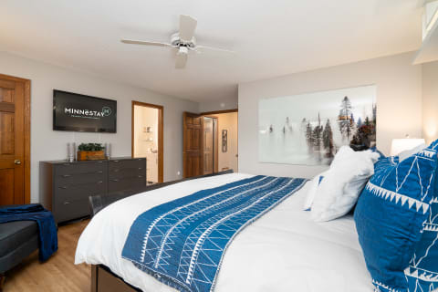 A cozy bedroom with white bedding and a blue quilt, decorated with a TV and a large artwork.