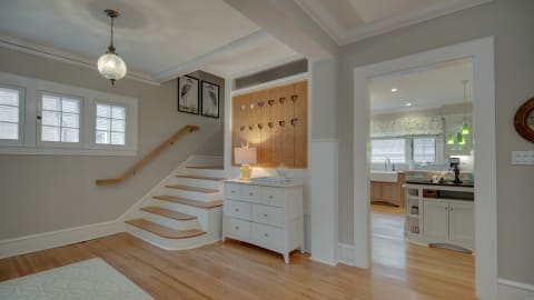 A stylish entryway featuring hardwood floors, a wooden staircase, a white dresser, and a glimpse into a modern kitchen.