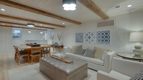 Cozy living space featuring a light-colored sofa, a silver tufted ottoman, and a wooden dining table beneath exposed beams.