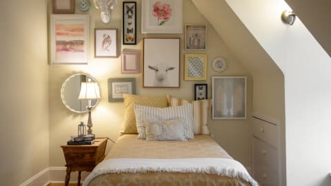 A cozy attic bedroom featuring a bed with decorative pillows, a wooden nightstand with books, and a gallery wall of framed artwork.