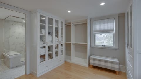 Walk-in closet with glass-front cabinets, a seating area, and a view into an ensuite bathroom.
