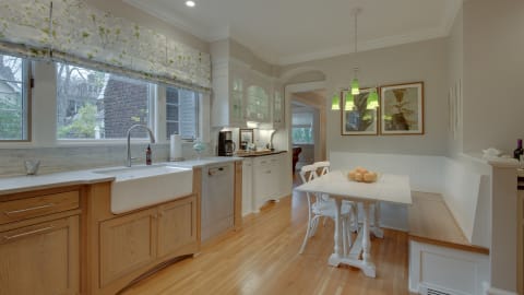Bright and inviting kitchen featuring a farmhouse sink, wooden breakfast nook, green pendant lights, and floral curtains.