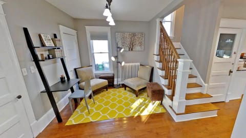 A bright living room with hardwood floors, two chairs, a small ottoman, and a black desk, enhanced by yellow accents.