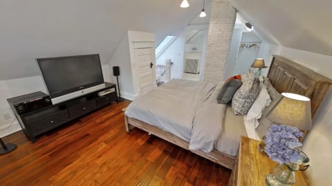 A cozy attic bedroom featuring rustic wooden floors, a plush bed with decorative pillows, and modern furnishings, illuminated by warm lighting.
