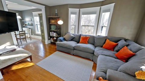 Cozy living room with gray sectional sofa, orange cushions, hardwood floors, and natural light through bay windows.
