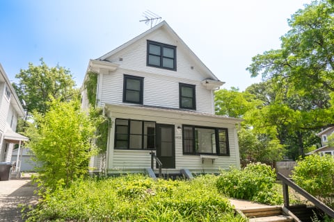 A white Victorian house with black-framed windows and a green door, set in a lush garden with trees around.