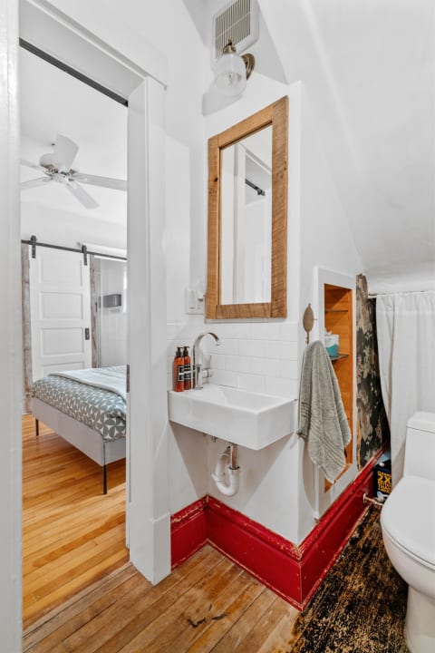 A contemporary bathroom with a floating sink, wooden mirror, and glimpse of a stylishly decorated bedroom.