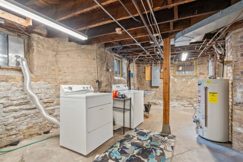 A rustic laundry room with a washer, dryer, sink, and colorful rug in a stone-walled basement.