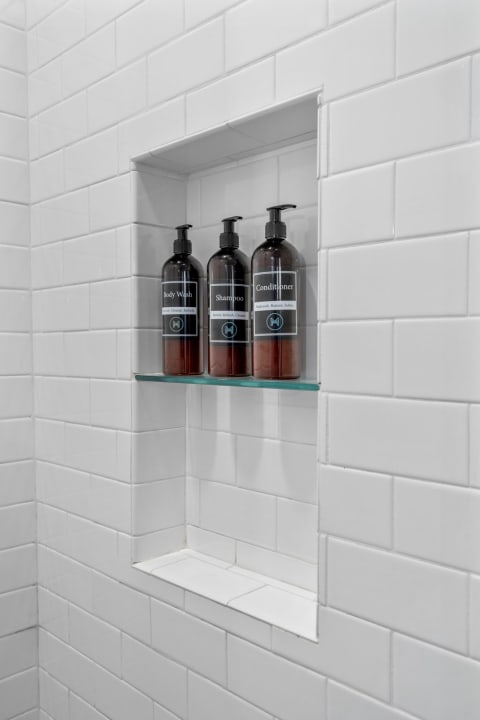 Black bottles of body wash, shampoo, and conditioner arranged in a shower niche against white tiled walls.