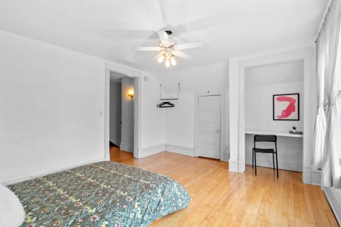A spacious bedroom with hardwood floors, a floral quilted bed, a workspace, and bright lighting.
