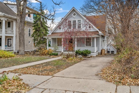 Victorian house with a gabled roof and porch in a residential area, surrounded by trees and autumn leaves.