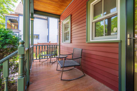 A covered porch featuring comfortable rocking chairs and a small table, set against a colorful wooden wall and lush greenery.