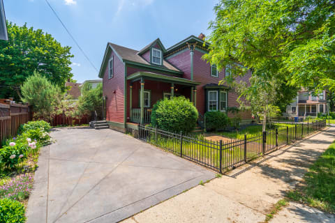 Victorian-style house with red exterior and front porch, surrounded by landscaped gardens.