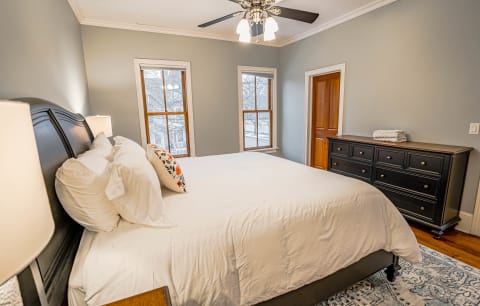 A cozy bedroom featuring a large bed with plush white bedding, floral accent pillow, and two windows that let in natural light.